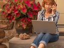 Woman on porch using laptop and phone beside red potted leaves; text "SENIOR PLANET"