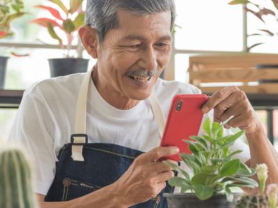 Older man in apron using a red smartphone while tending potted plants; text "SENIOR PLANET"
