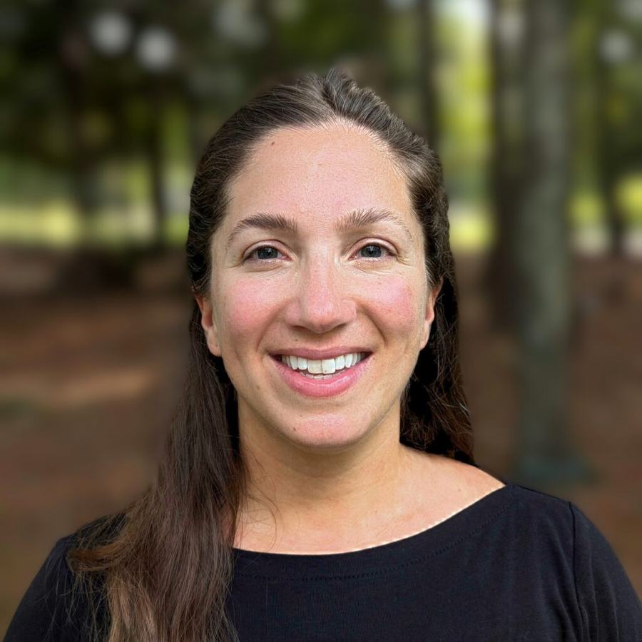 Smiling woman facing camera, close-up headshot with blurred trees in background