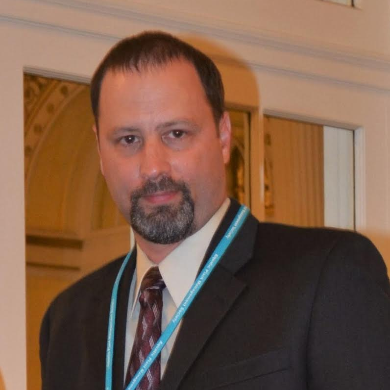 Man in dark suit and patterned tie wearing a blue lanyard, facing camera