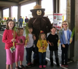 Six children holding plants beside a person in a bear costume wearing a straw hat