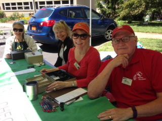Four volunteers sit at an outdoor registration table, smiling at the camera, with event materials and a car visible in the background.