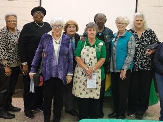 Group of 12 older adults standing side-by-side in a community room, one wearing an apron.