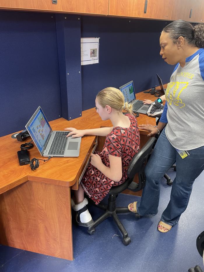A woman looks on as a girl creates a video game on a laptop.