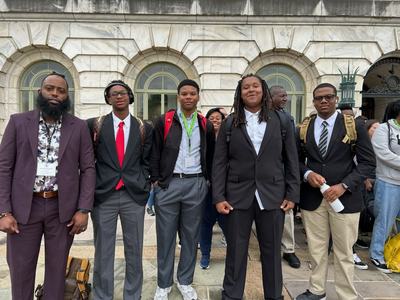 Group of young men in suits and backpacks standing outside a marble building