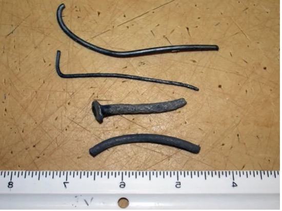 Four corroded bent nails on a wooden surface beside a ruler with inch markings.