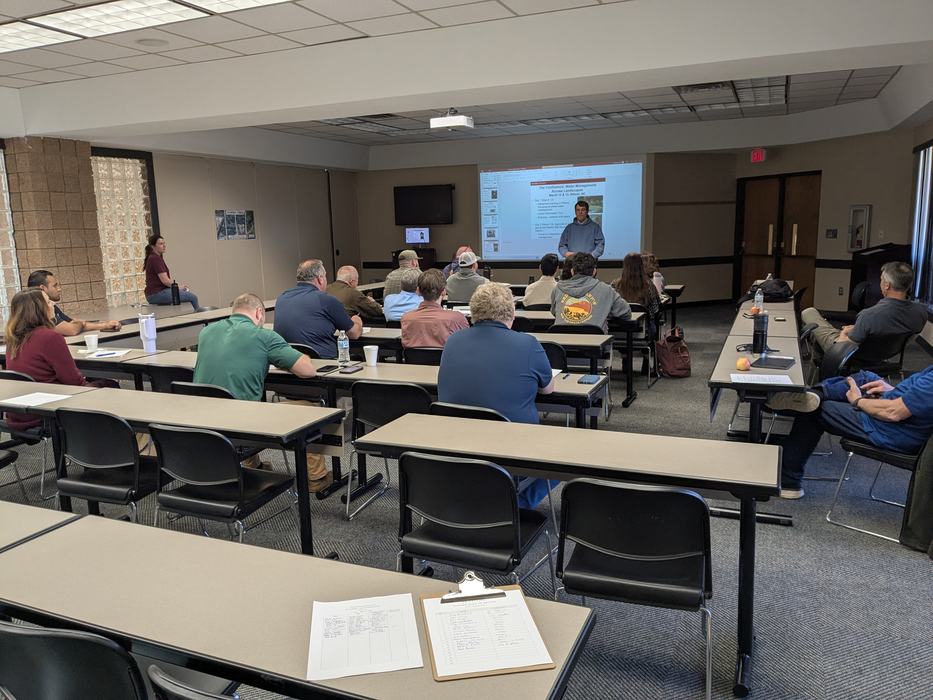 Classroom lecture with presenter at front projecting a slide and attendees seated facing forward