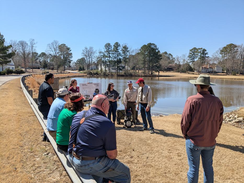 People gathered by a pond listening to presenters standing near a display board