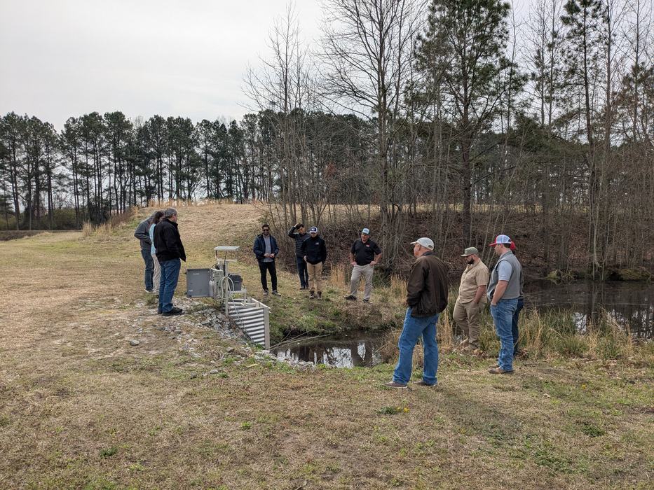 Group of people standing by pond inspecting water-monitoring equipment on the bank