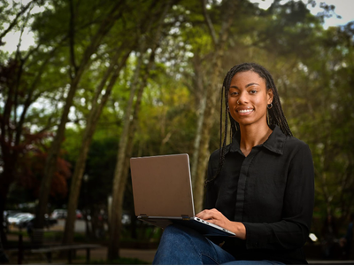 Young woman sitting outdoors using a laptop on her lap in a tree-lined park