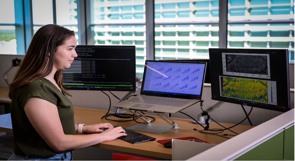 Woman at standing desk typing with laptop and two monitors showing code and data plots