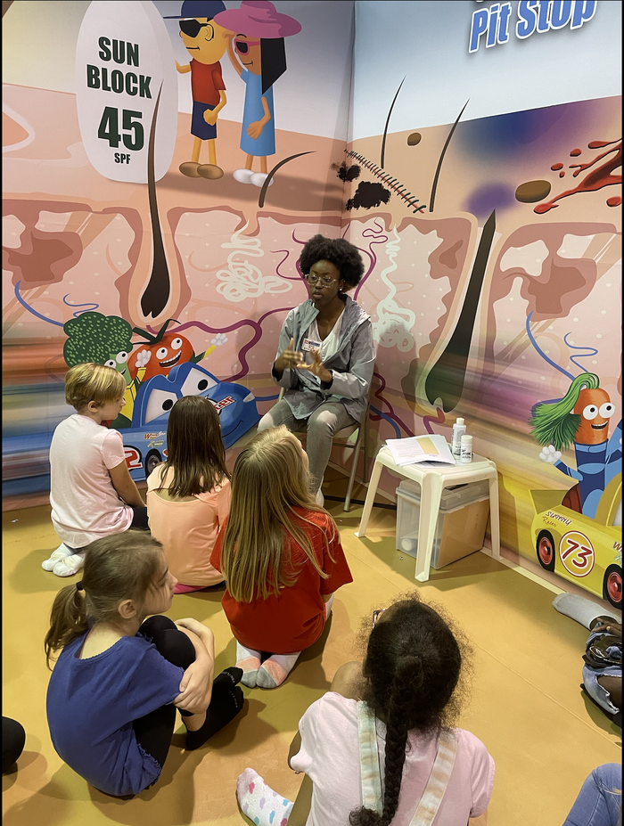 Woman speaking to children in mural booth, "SUN BLOCK 45 SPF" sign visible