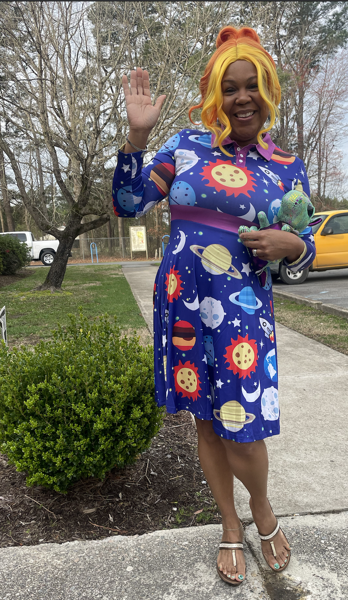 Person waving in blue space-pattern dress with orange-yellow hair, holding a plush turtle