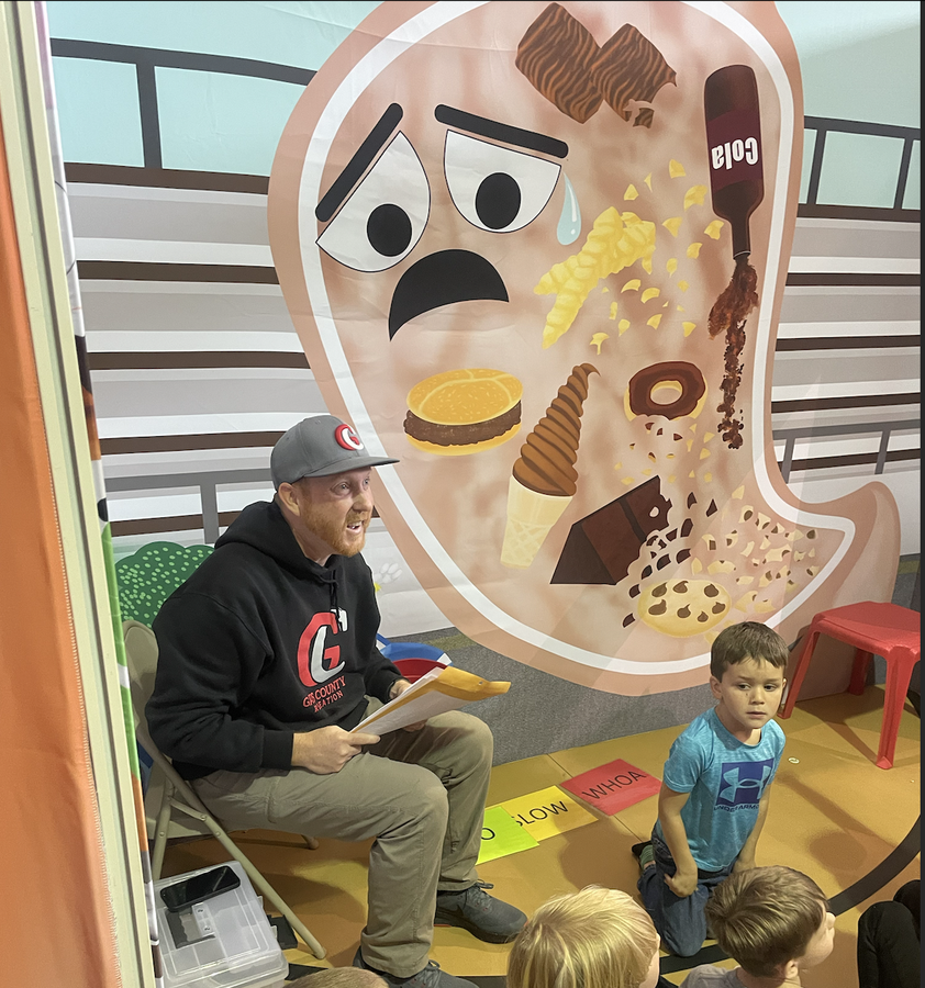 Man reading to children in front of a poster showing a stomach illustration with a tear and junk food