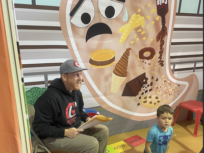 Man reading to children in front of a poster showing a stomach illustration with a tear and junk food