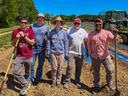 Five farm workers standing on a planted field with shovels and a tractor behind