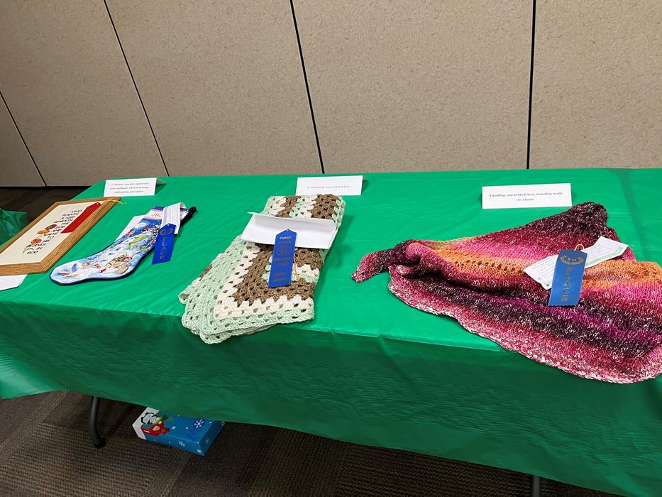Table with knitted and crocheted items on green tablecloth with blue prize ribbons