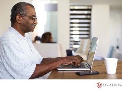 Older man typing on laptop at table with phone and mug; text "SENIOR PLANET"