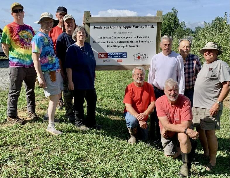 Group of people, including Master Pomologists, posing by a sign for the Henderson County Apple Variety Block.