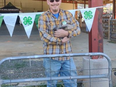 Man in plaid shirt holding a rabbit in front of 4‑H clover bunting at a fair