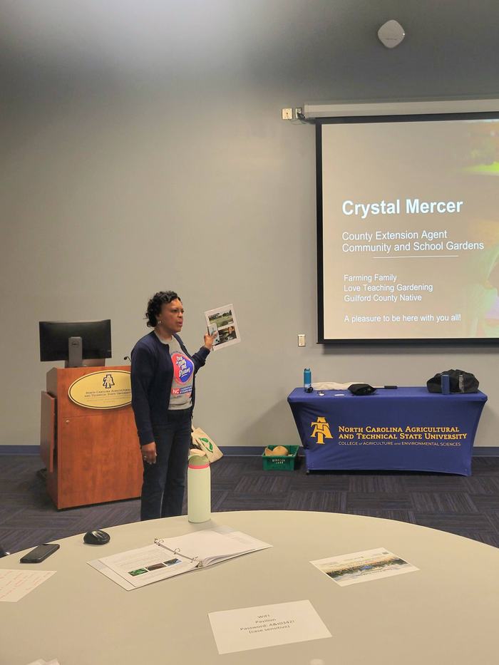 Crystal is standing to the left of her presentation on a projector while doing her lecture. She is gesturing towards the presentation, which currently shows her role in Extension.