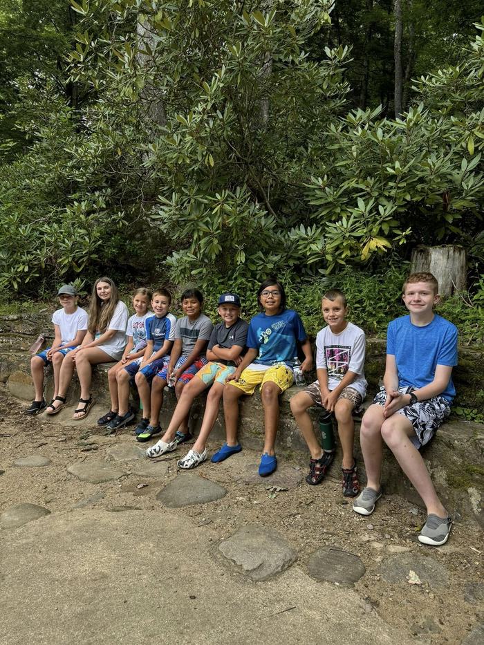 Ten children sitting on a low stone wall in front of dense green bushes on a dirt path