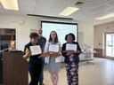 Three women stand with certificates and NC wooden awards.