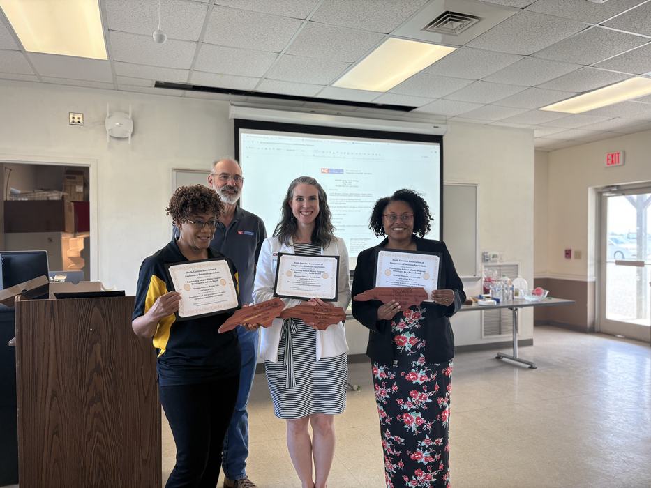 Three women stand with certificates and NC wooden awards. 