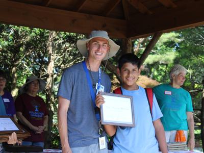 Man in sun hat standing with boy holding a framed certificate under a pavilion