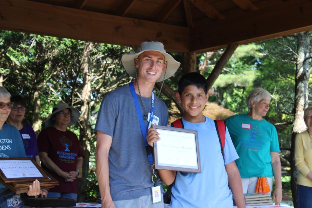 Man in sun hat standing with boy holding a framed certificate under a pavilion