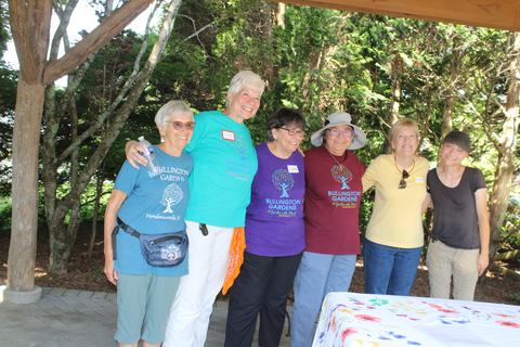 Six women posing arm‑in‑arm under a pavilion, wearing "Bullington Gardens" shirts