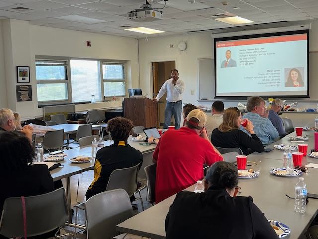 Room of people at long tables look to presenter at the front of the room. 