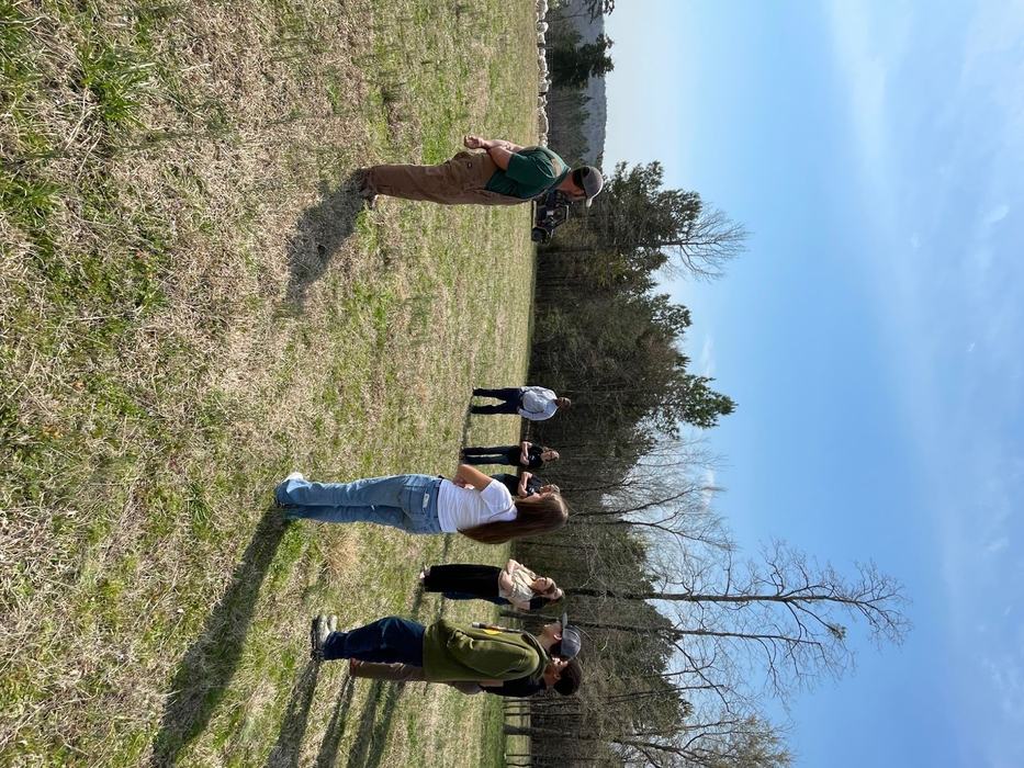 People form a semi-circle around a man in overalls in an open field.