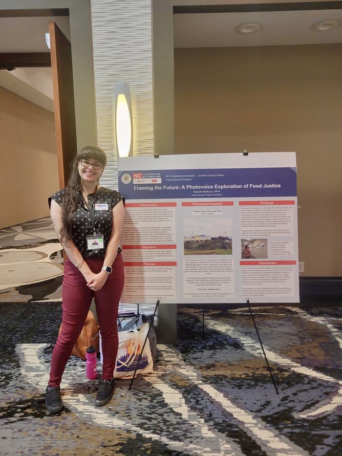 Melodie is posing to the left of her poster in a black polka-dot shirt and red pants.