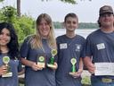 Four teens in grey t-shirts holding their 1st place trophies pose for a picture with Lake James in the background.