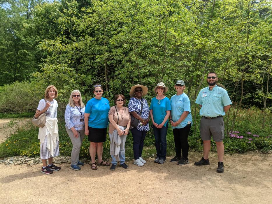 Nine adults standing side-by-side on a park path in front of dense green trees