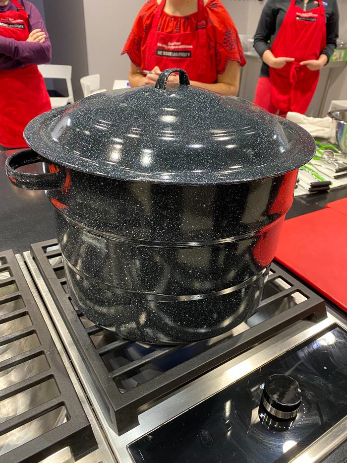 Black speckled enamel stockpot with lid on a gas stove, people in red aprons in background