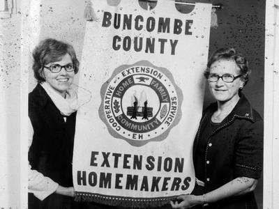 Black and White photo of two women holding a banner saying Buncombe County Extension Homemakers during the 1970s.
