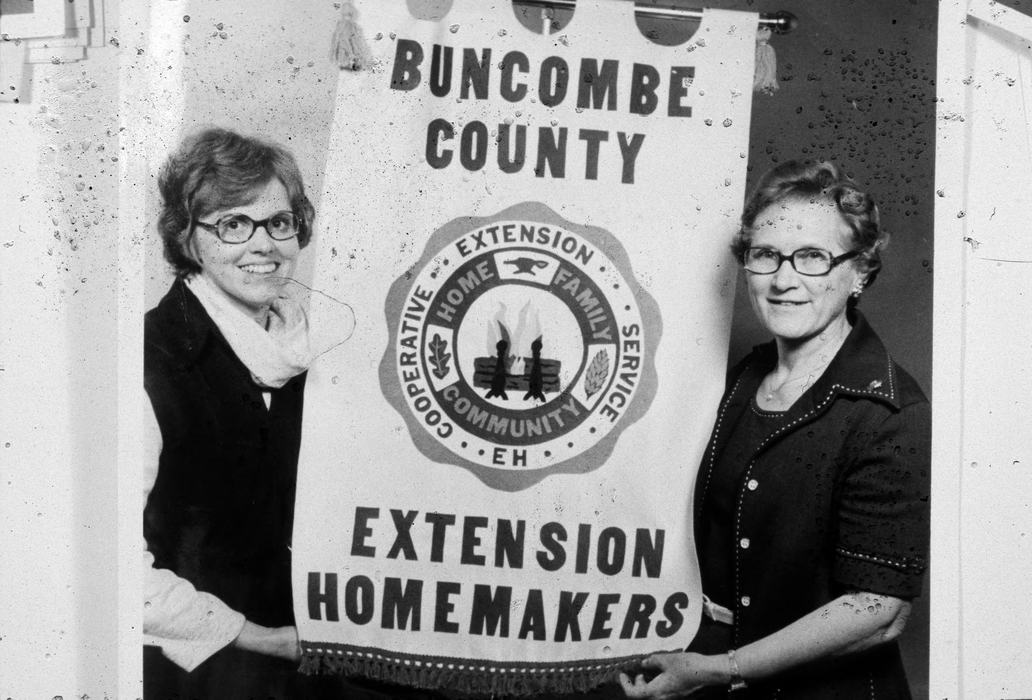 Black and White photo of two women holding a banner saying Buncombe County Extension Homemakers during the 1970s.