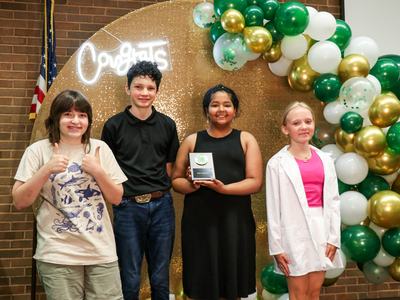 Four children standing in front of a gold backdrop and green balloons; center girl holds a 4‑H award plaque.