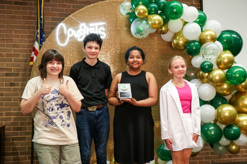 Four children standing in front of a gold backdrop and green balloons; center girl holds a 4‑H award plaque.