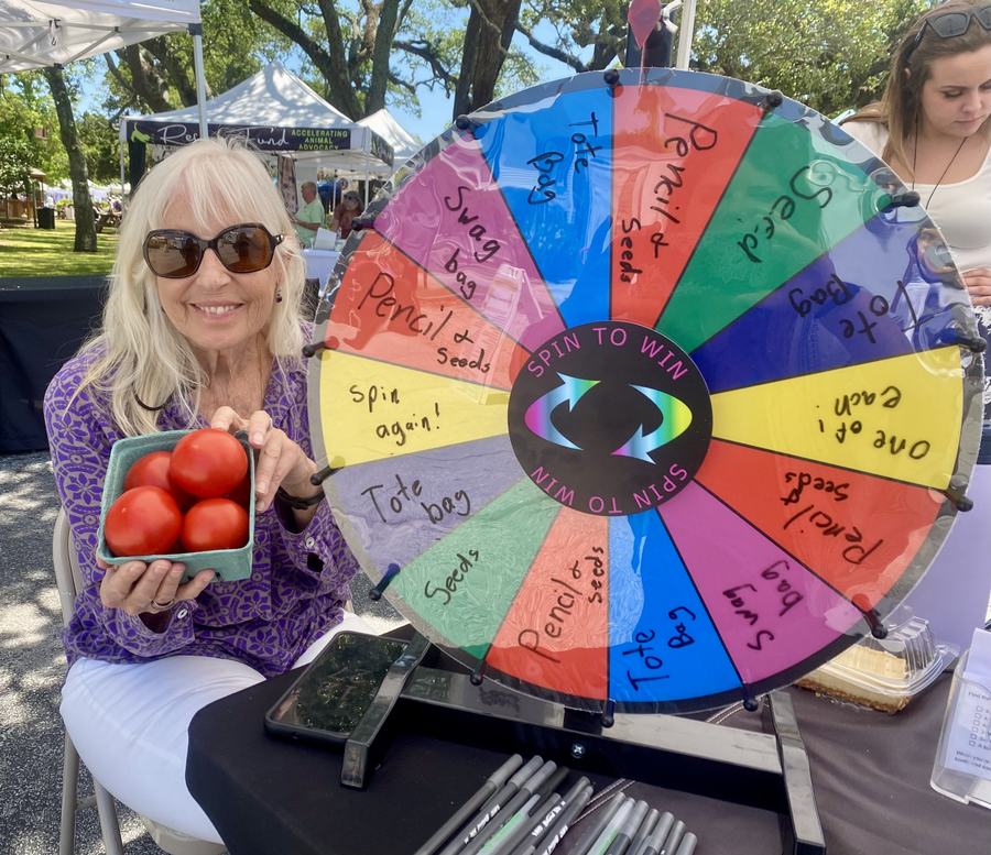 A woman holds a carton of fresh tomatoes beside a spin to win prize wheel.