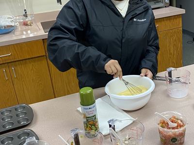 Person whisking eggs in a bowl on a counter with measuring cups of chopped vegetables and cheese