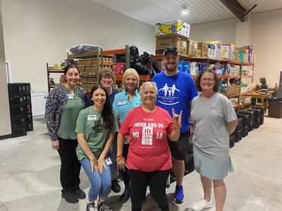 Eight volunteers posing in a warehouse food pantry in front of stocked shelves