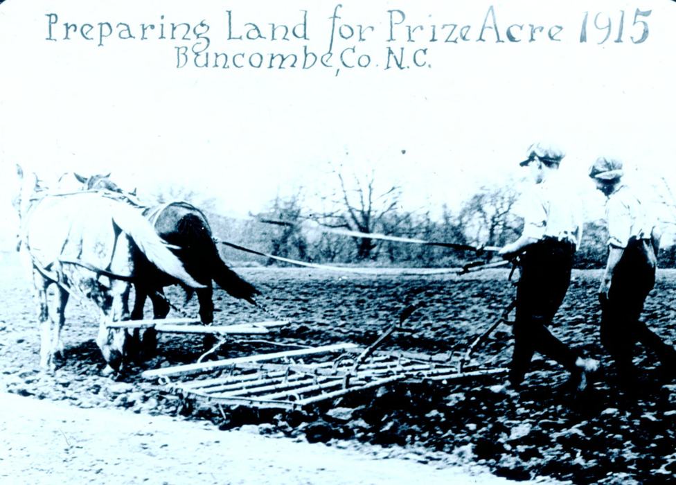 Black and white photo from 1915 of two boys working in a field behind one white horse and one dark horse pulling a farming implement. 