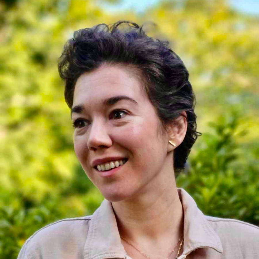 Person with short dark hair smiling, wearing light jacket and gold earrings outdoors