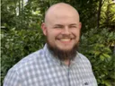 Bald man with beard smiling, wearing checkered shirt in front of leafy background