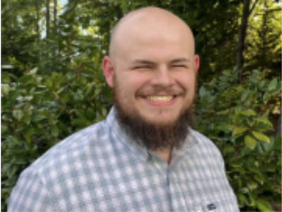 Bald man with beard smiling, wearing checkered shirt in front of leafy background
