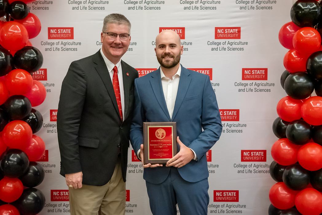 Two men stand in front of an NC State University College of Agriculture and Life Sciences backdrop; one holds a red and gold award plaque while both smile for the camera.