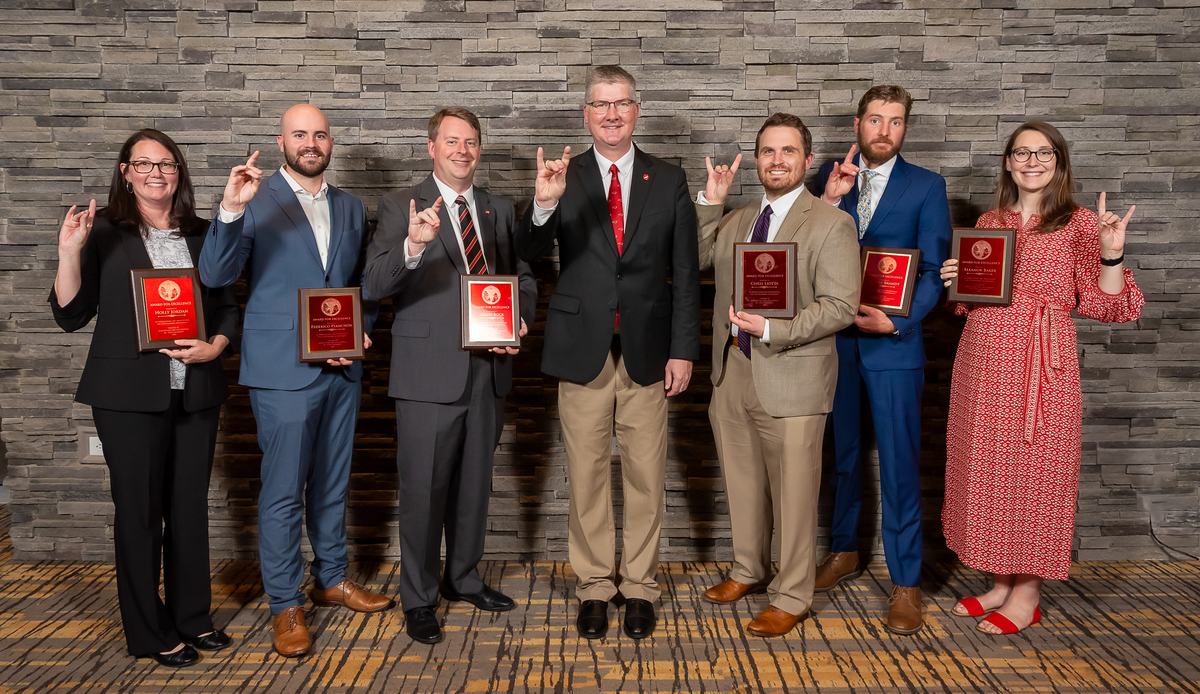 Seven award recipients pose in front of a stone wall, each holding a plaque and making the NC State “wolf hand” gesture, dressed in business attire.